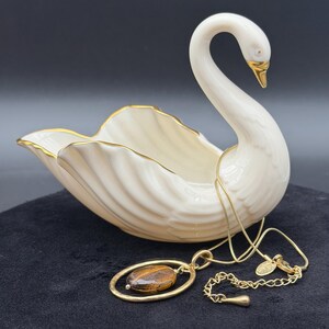 May include: A cream-colored ceramic swan dish with gold-painted trim on the wings and beak. A gold-tone necklace with a thin chain and an oval pendant featuring a brown and black tiger eye stone rests in front of the swan figurine.