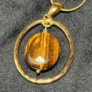 May include: A gold-toned pendant necklace featuring a polished oval tiger's eye gemstone suspended within a circular gold-toned frame. The gemstone displays characteristic brown and golden chatoyant bands, and the pendant hangs from a matching chain.