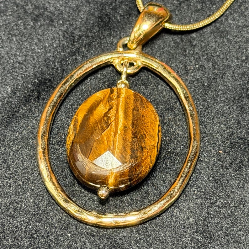 May include: A gold-toned pendant necklace featuring a polished oval tiger's eye gemstone suspended within a circular gold-toned frame. The gemstone displays characteristic brown and golden chatoyant bands, and the pendant hangs from a matching chain.