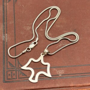 May include: A silver-toned necklace with a unique pendant. The pendant has an open, abstract design. The chain is a snake chain style, and the clasp is visible. The necklace rests on a dark red surface.