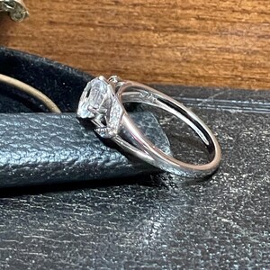 May include: A silver ring with a large, clear gemstone and smaller accent stones. The ring's band is smooth and polished, and the setting has intricate details. The ring is displayed on a dark surface.