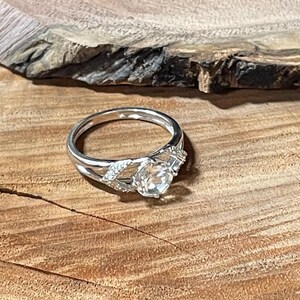 May include: A silver ring with a clear, round gemstone as the focal point. The ring features a split band design with smaller accent stones on either side of the main stone. The ring is set against a wooden surface.