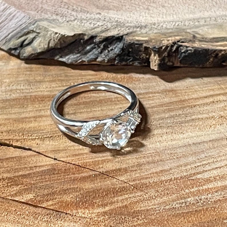 May include: A silver ring with a clear, round gemstone as the focal point. The ring features a split band design with smaller accent stones on either side of the main stone. The ring is set against a wooden surface.