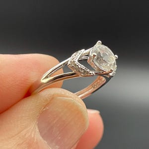 May include: A silver ring with a large, clear, oval-cut gemstone. The ring features a split shank design and small accent stones on the shoulders. The ring is held by a person's fingers against a dark background.
