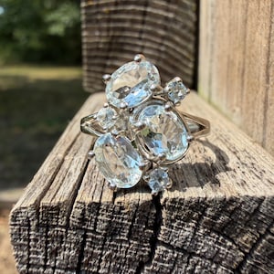 May include: A silver-toned ring featuring a cluster of five faceted, light blue oval and round gemstones. The stones are arranged in a tiered formation, with three larger oval stones and two smaller round stones set in a metallic band structure.