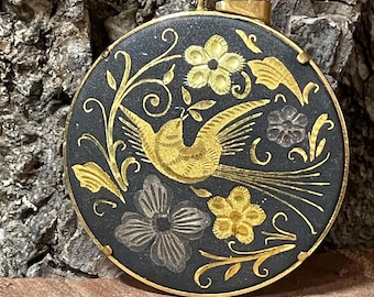 May include: A round pendant with a black background and a gold-colored bird in flight, surrounded by gold and silver floral designs. The pendant has a gold-colored frame and a loop at the top for a chain.