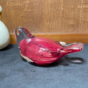 May include: A decorative glass bird figurine. The bird is primarily a deep red color with clear glass wings and head. The red glass has a swirled pattern and small air bubbles. The bird is resting on a dark surface.