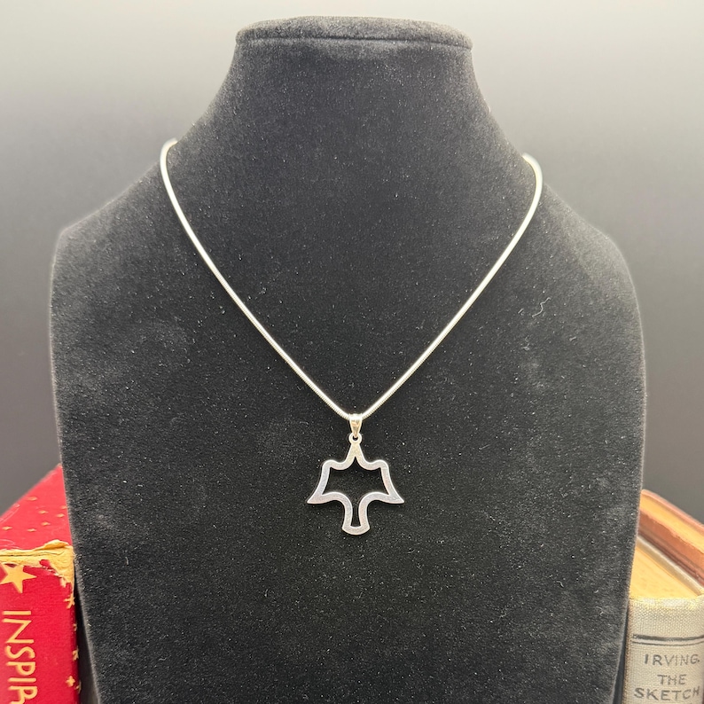 May include: A silver-toned pendant necklace featuring a stylized dove design. The pendant is a cutout shape, and the necklace is displayed on a black mannequin. The dove pendant hangs from a delicate silver chain.
