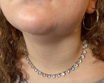 May include: A silver-toned necklace with clear, round gemstones. The necklace is worn around the neck and is paired with silver hoop earrings. The person is wearing a gray shirt.