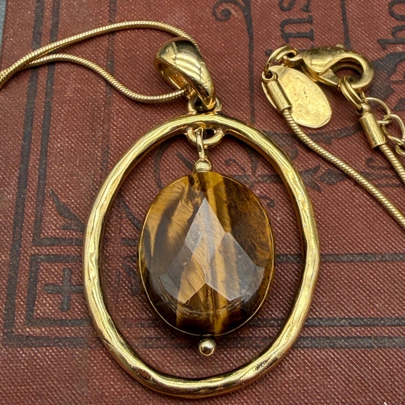 May include: A gold-tone necklace featuring a faceted oval tiger's eye gemstone suspended within a textured, hammered-finish gold-tone oval frame. The pendant hangs from a thin gold-tone snake chain with a lobster claw clasp and a small metal tag.