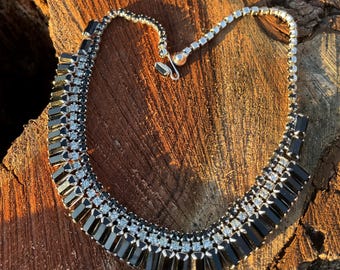 May include: A silver-toned necklace with black rectangular and clear square-cut stones. The necklace is set on a wooden surface, showcasing its design and sparkle. The clasp is a hook and eye closure.