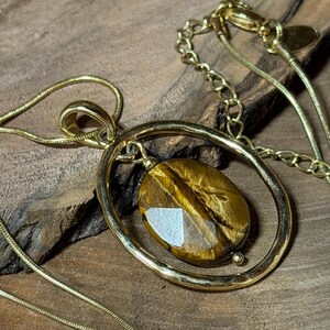 May include: A gold-toned necklace featuring a polished oval frame pendant that holds a faceted, golden-brown tiger's eye gemstone. The pendant is suspended from a delicate gold-toned snake chain that includes an adjustable link extender.