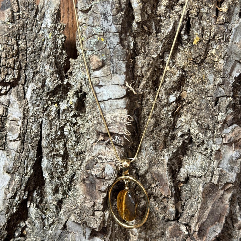 May include: A gold-toned necklace featuring a thin chain and a circular pendant. The pendant contains a single, smooth, oval-shaped tiger's eye gemstone suspended in the center of a polished gold-colored metal ring, creating a layered appearance.