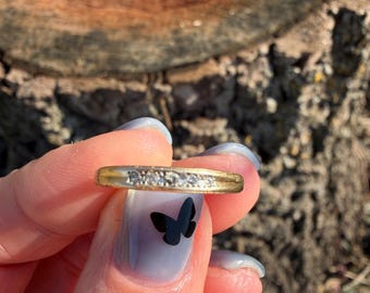 May include: A gold-toned ring with a row of small, clear stones. The ring is held up against a blurred background of a tree trunk. The ring is a classic design, suitable for everyday wear or special occasions.