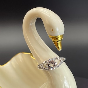 May include: A silver ring with a central, light-colored gemstone, set within a diamond-shaped frame, is displayed on a white swan figurine. The swan has a gold beak and accents, with a smooth, glossy finish.