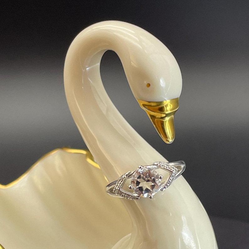 May include: A silver ring with a central, light-colored gemstone, set within a diamond-shaped frame, is displayed on a white swan figurine. The swan has a gold beak and accents, with a smooth, glossy finish.