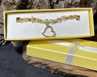 May include: A gold-toned bracelet with a heart pendant, presented in a yellow gift box with a white ribbon. The bracelet features a series of small, decorative links and clear gemstones. The heart pendant is outlined with a delicate design and accented with small gemstones.