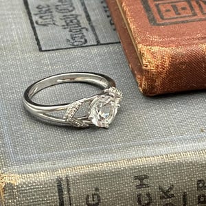 May include: A silver ring with a large, clear, round gemstone as the centerpiece. The band splits into a decorative design on either side of the gemstone, with small accent stones. The ring is set against a vintage book.
