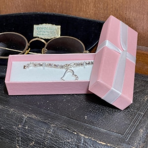 May include: A silver bracelet with a heart charm rests in a pink gift box tied with a white ribbon. The bracelet features rectangular and round embellishments. A pair of gold-rimmed glasses and a black case are in the background.