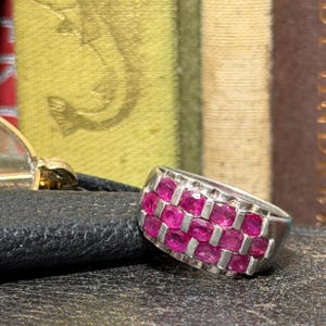 May include: A silver-toned ring featuring two horizontal rows of small, round, vibrant pink gemstones set in a basket-weave pattern. The band is wide and metallic, showcasing a repeating grid design that holds each individual stone securely in place.
