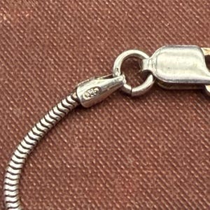 May include: Close-up of a silver necklace clasp and chain. The chain has a snake-like design, and the clasp is connected to a small, rounded ring. The clasp is stamped with "925".
