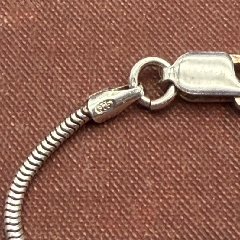May include: Close-up of a silver necklace clasp and chain. The chain has a snake-like design, and the clasp is connected to a small, rounded ring. The clasp is stamped with "925".