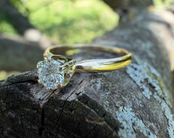 May include: A gold engagement ring with a large, round-cut diamond. The ring is set on a textured, wooden surface with a blurred green background. The diamond sparkles, reflecting light.