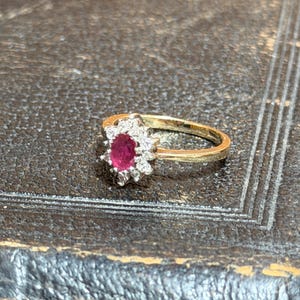 May include: A gold ring featuring a central oval ruby surrounded by a halo of small, clear gemstones. The ring's band is a simple, polished gold. The ring is set against a dark, textured surface.