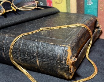 May include: A gold-colored chain necklace with a textured design lies on a worn, black leather-bound book. The necklace is draped across the book's surface, with a clasp visible. Vintage eyeglasses in a black case are in the background.