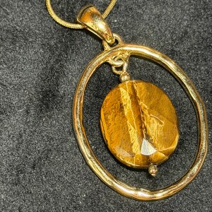 May include: A gold-toned pendant necklace featuring a large, oval-shaped tiger's eye gemstone suspended within a textured, irregular gold-toned circular frame. The pendant hangs from a matching gold-toned snake chain, showcasing a polished finish.