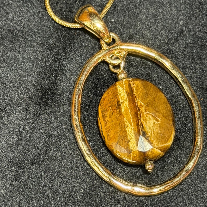 May include: A gold-toned pendant necklace featuring a large, oval-shaped tiger's eye gemstone suspended within a textured, irregular gold-toned circular frame. The pendant hangs from a matching gold-toned snake chain, showcasing a polished finish.