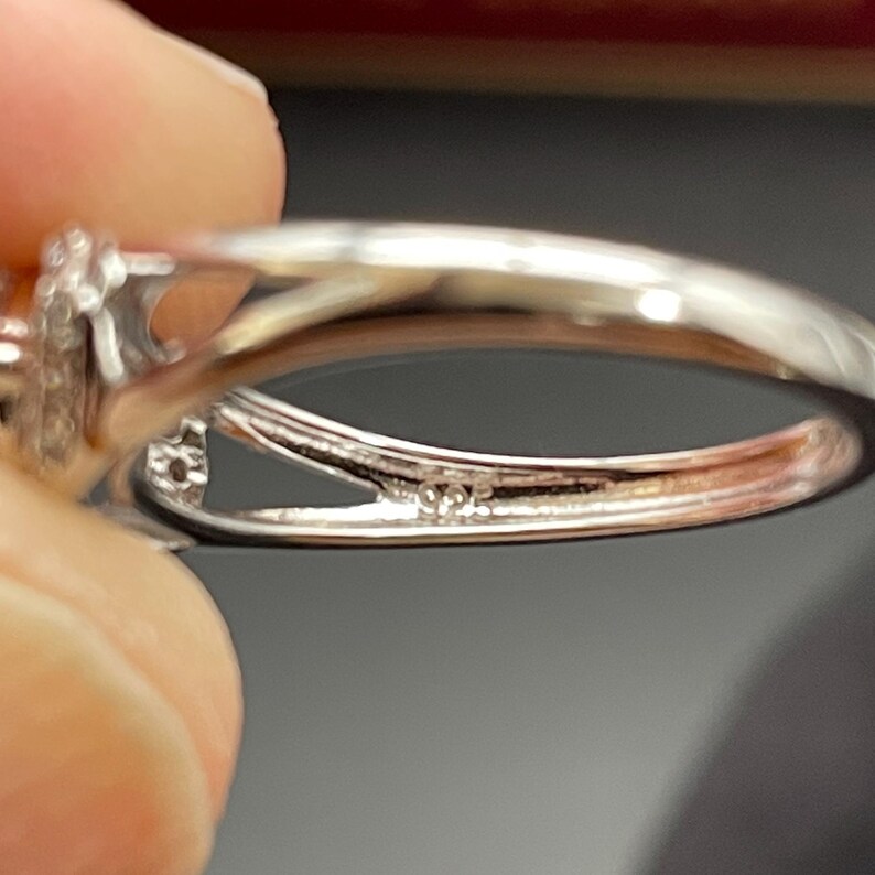 May include: Close-up of a silver ring with a split shank design. The ring has a small, decorative element with what appears to be small stones. The inside of the ring is stamped with "925".