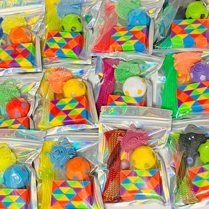 May include: A collection of clear, sealed bags filled with colorful fidget toys. Each bag contains a variety of items, including textured balls, geometric puzzles, and stretchy tubes. The toys are in various bright colors, including red, yellow, blue, and green.