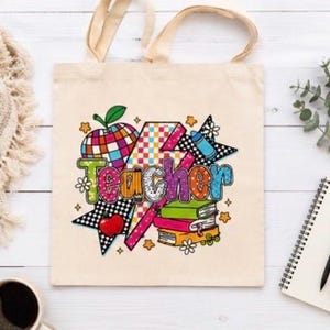 Può includere: Borsa tote beige con un design grafico colorato con la parola "Teacher" in grandi lettere multicolori. Il design include una mela, libri, stelle e altri elementi a tema scolastico. La borsa ha manici color crema.