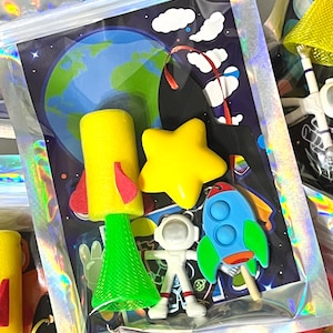 May include: A clear plastic bag filled with space-themed charms. The charms include a yellow star, a rocket, an astronaut, and a yellow and red space shuttle. The background features a planet Earth and a starry sky.