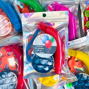 K&ouml;nnte beinhalten: Eine transparente Plastikt&uuml;te gef&uuml;llt mit bunten Fidget-Spielzeugen, darunter ein Pop-it, ein Fidget Spinner und ein biegsamer Strohhalm. Die T&uuml;te hat ein Etikett mit der Aufschrift "Danke, dass du mit mir gefeiert hast! - Hunter".