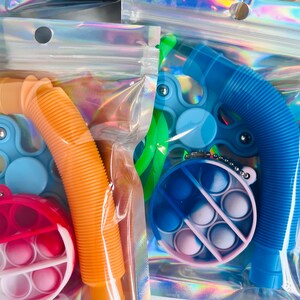 K&ouml;nnte beinhalten: Sortiment an Fidget-Spielzeug in einzelnen durchsichtigen Plastikt&uuml;ten. Das Spielzeug umfasst einen blauen und gr&uuml;nen Fidget Spinner, ein blaues Pop-R&ouml;hrchen, ein orangefarbenes Pop-R&ouml;hrchen, ein blaues und pinkfarbenes Pop It und ein rotes und wei&szlig;es Pop It.