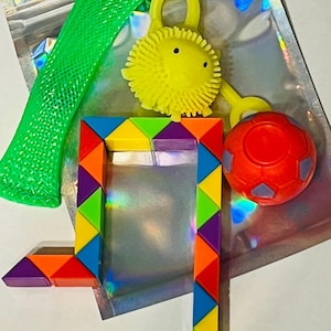 May include: A collection of colorful sensory toys, including a green mesh tube, a yellow spiky ball with a loop, a red ball, and a geometric puzzle. The toys are arranged on a shiny, iridescent surface.
