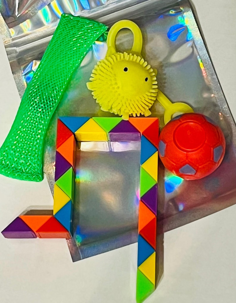 May include: A collection of colorful sensory toys, including a green mesh tube, a yellow spiky ball with a loop, a red ball, and a geometric puzzle. The toys are arranged on a shiny, iridescent surface.