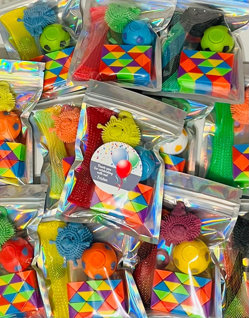 May include: A collection of clear plastic bags filled with colorful toys. Each bag contains a variety of items, including spiky balls, rainbow-colored puzzles, and other small toys. Some bags have a sticker that says "Thank you for celebrating with me! -Tristen."