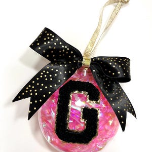 May include: Clear Christmas ornament filled with pink confetti and a black letter "G" with gold trim. A black ribbon with gold polka dots is tied in a bow at the top, and a gold ribbon is used for hanging.