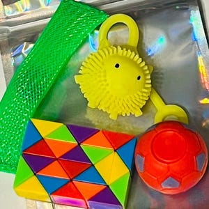 May include: A collection of colorful fidget toys, including a green mesh tube, a yellow spiky ball with a loop, a geometric puzzle, and a red soccer ball-shaped toy. The toys are arranged on a reflective surface.