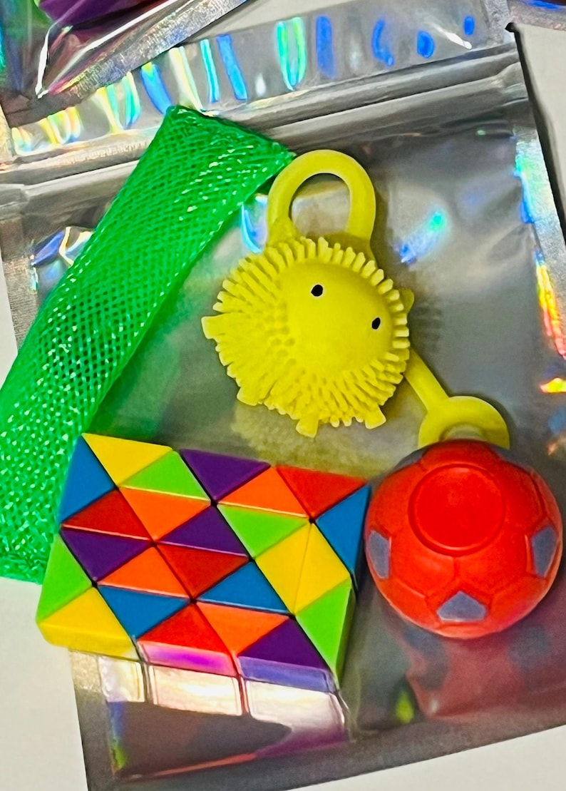 May include: A collection of colorful fidget toys, including a green mesh tube, a yellow spiky ball with a loop, a geometric puzzle, and a red soccer ball-shaped toy. The toys are arranged on a reflective surface.