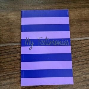 May include: A notebook with a purple and blue striped cover. The text "My Testimonies" is written in cursive across the front. The notebook is lying on a wooden surface.