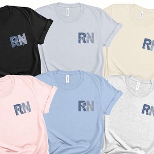 Puede incluir: Cinco camisetas de diferentes colores con el texto "RN" y un gr&aacute;fico de coraz&oacute;n estilizado. Las camisetas son todas de manga corta y tienen cuello redondo.