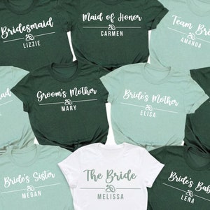 May include: A collection of ten short-sleeved t-shirts in dark green, light green, and white. Each shirt features white text with titles like Bridesmaid, Maid of Honor, Team Bride, Bride Squad, Groom's Mother, Bride's Mother, and The Bride.
