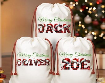 May include: Three white Christmas gift bags with red drawstrings. Each bag has a name in red and black plaid letters with Santa hats above the letters. The bags read "Merry Christmas" in green script above the names "Jack", "Oliver", and "Zoe".