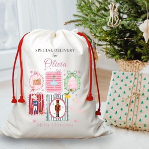 May include: A cream-colored Christmas gift bag with red drawstrings and tassels. The bag is decorated with colorful illustrations and the text "Special Delivery for Olivia" and "Merry Christmas". A wrapped gift and Christmas tree are in the background.