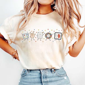 May include: A cream-colored short-sleeve t-shirt featuring a graphic print of five anthropomorphic school-themed characters including a square grid, a hand making a sign, a calculator, a colorful wreath, and a timer, all surrounded by small stars.