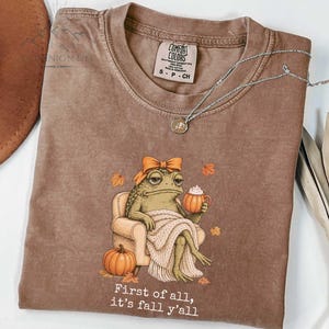 May include: A brown short-sleeve t-shirt featuring a graphic of a toad wearing an orange bow, sitting in a chair with a blanket, holding a pumpkin mug. The text reads, First of all, it's fall y'all. The shirt has a Comfort Colors brand label.